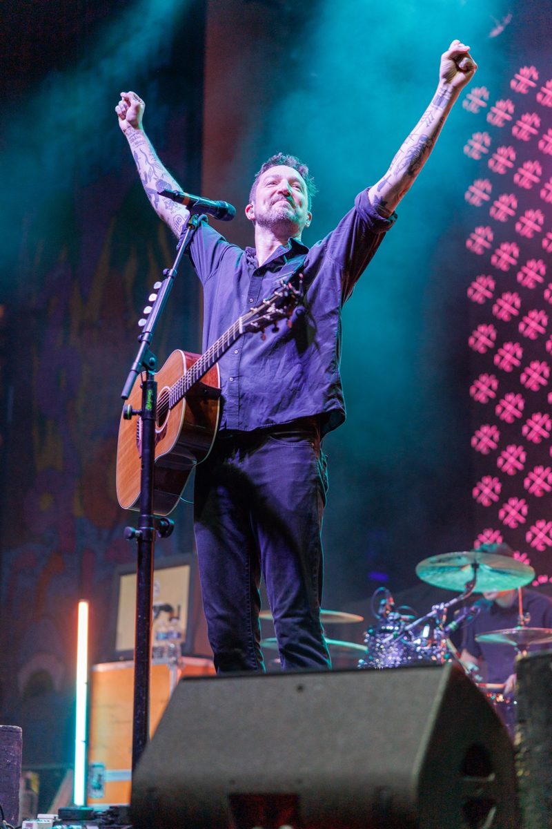 Frank Turner with guitar strapped on, both arms raised in the air on stage